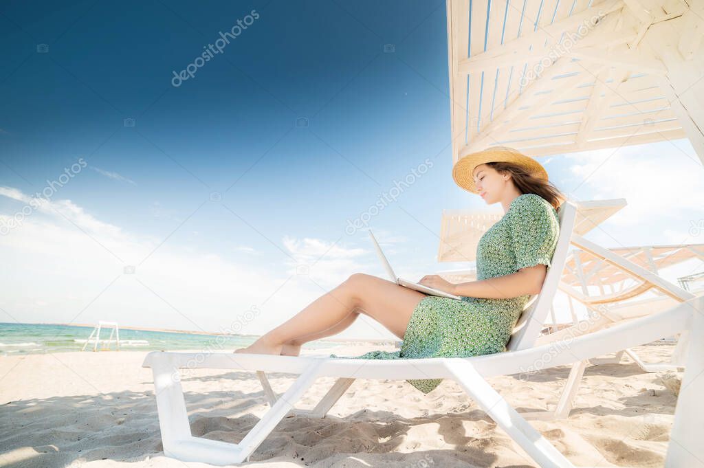 Lovely young caucasian woman freelancer in a green dress and straw hat on a schizlong under an umbrella works on her laptop. Remote work on the seaside success