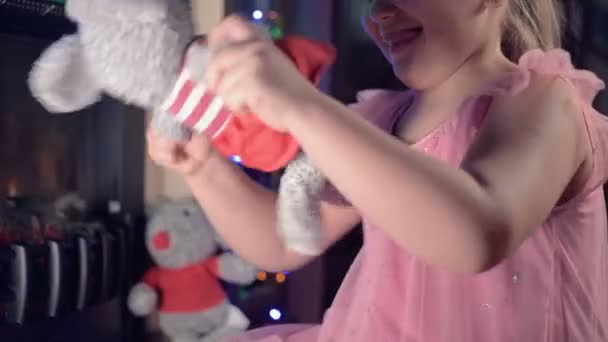 Lite ensam glad fet flicka leker med en mjuk leksak råtta på bakgrunden av en elektrisk öppen spis och blinkande nyårsljus på julafton — Stockvideo