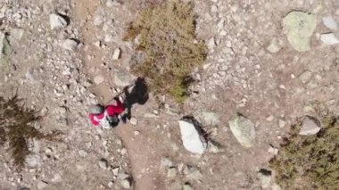Şapkalı ve şortlu bir adamın havadan görünüşü ve sırt çantasıyla dağlardaki dağ vadisi boyunca yürüyüş direkleri ile yürüyor.