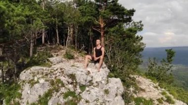 Uzun saçlı, siyah tişörtlü ve şortlu zayıf bir adam uçurumun kenarında oturur ve etrafındaki orman ve dağlara bakar. Hava görünümü
