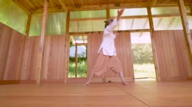 Uzun saçlı, açık renk elbiseli beyaz bir adam yaz aylarında tahta bir çalışma odasında qigong tai chi yapıyor. Yavaş hareketler uyum ve sakinlik