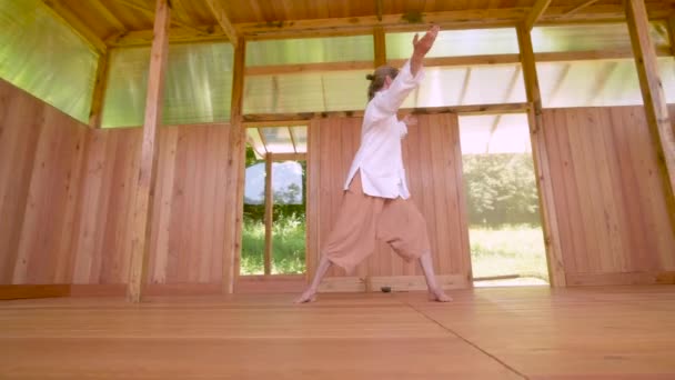 Een langharige blanke man in lichte en losse kleding oefent 's zomers qigong tai chi in een houten oefenruimte. Langzame bewegingen harmonie en rust — Stockvideo