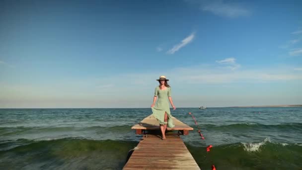 An attractive Caucasian girl in a green summer dress and a straw hat walks along the wooden pier of the sea coast in sunny weather. Rest at the sea. Travel video — Stock Video