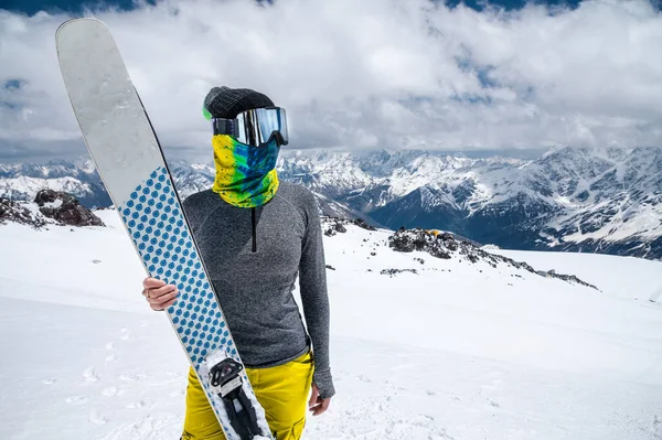 An attractive slender woman skier stands without a jacket high in the mountains and holds her skis against the background of high snow-capped peaks and clouds — Stock Photo, Image