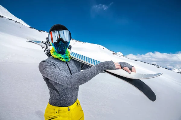 En attraktiv smal kvinnlig skidåkare står utan jacka högt uppe i bergen och håller sina skidor på axeln mot bakgrund av höga snötäckta toppar och moln — Stockfoto