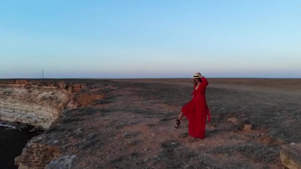 Widok z lotu ptaka młodej kobiety w czerwonej sukience i słomkowym kapeluszu spaceruje wzdłuż krawędzi klifu nad morzem samotnie o zachodzie słońca — Wideo stockowe