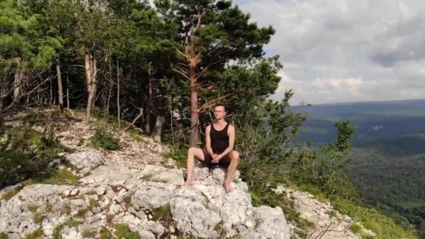 Un homme mince et aux cheveux longs portant un T-shirt noir et un short est assis sur le bord d'une falaise et regarde la forêt et les montagnes environnantes. Vue aérienne — Video