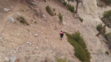 Şapkalı ve şortlu bir adamın havadan görünüşü ve sırt çantasıyla dağlardaki dağ vadisi boyunca yürüyüş direkleri ile yürüyor.