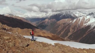 Sırt çantalı, güneş gözlüklü, yürüyüş direkleri olan genç bir adamın dağdan aşağı inişini diğer dağların ve geçitlerin arka planında karın yanında yürürken görüntüsü.