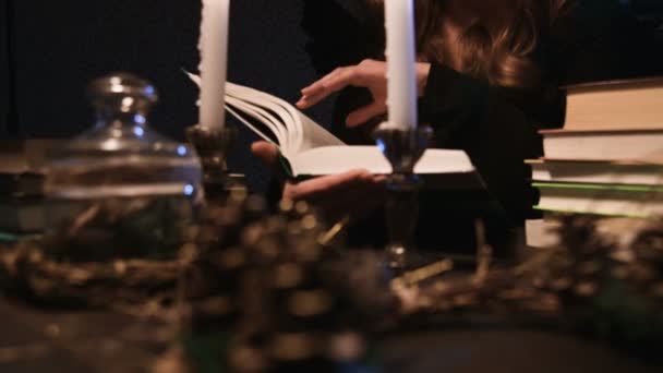 Een jonge vrouw met lang gouden haar in een kap gekleed in een heksenkostuum bij kaarslicht in een donkere kamer bladerend door een dik boek. Halloween — Stockvideo