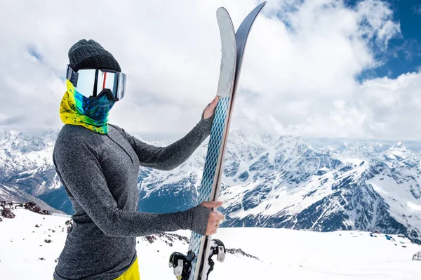 Μια ελκυστική λεπτή γυναίκα σκιέρ στέκεται χωρίς σακάκι ψηλά στα βουνά και κρατά τα σκι της με φόντο τις ψηλές χιονισμένες κορυφές και τα σύννεφα — Φωτογραφία Αρχείου