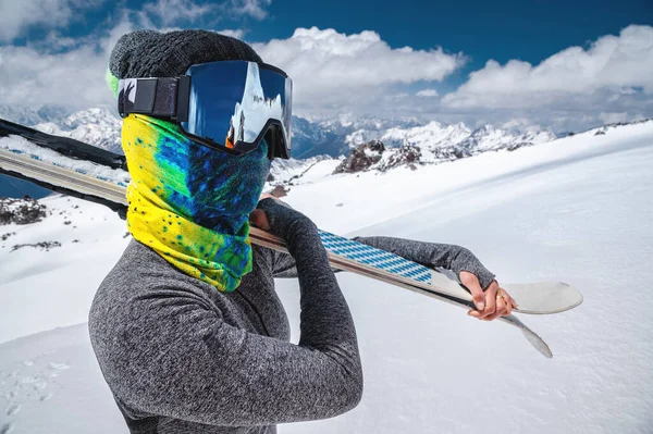 Eine attraktive, schlanke Skifahrerin steht ohne Jacke hoch in den Bergen und hält vor dem Hintergrund hoher schneebedeckter Gipfel und Wolken ihre Skier auf der Schulter. — Stockfoto