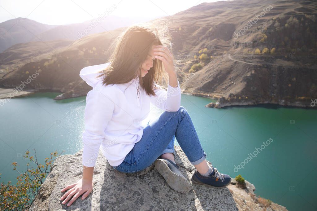Attractive young caucasian woman in white sweatshirt and jeans sits on a cliff on a cliff in front of a large mountain lake on a sunny day