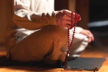 Meditasyon yapan bir keşişin erkek eli, mum ışığında karanlık bir odada kırmızı bir tespih tutar. Pratik ve aydınlanma