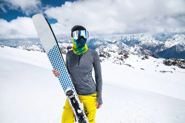 Eine attraktive, schlanke Skifahrerin steht ohne Jacke hoch in den Bergen und hält ihre Skier vor dem Hintergrund hoher schneebedeckter Gipfel und Wolken. — Stockfoto