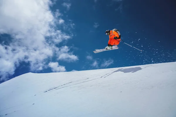Atleta professionista sciatore freerider in una tuta arancione con uno zaino vola in aria dopo aver saltato sui gal sullo sfondo del cielo blu e della neve — Foto Stock