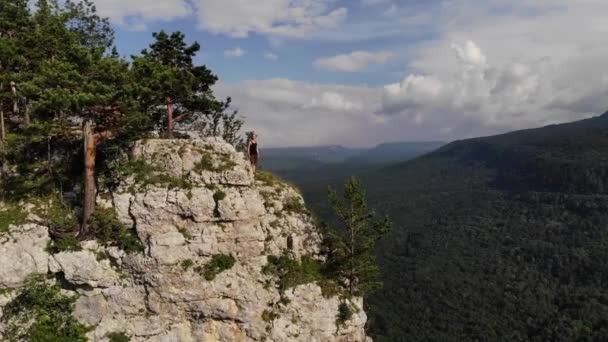 A long-haired, skinny, man in a black T-shirt and shorts stands on the edge of a cliff and looks out at the surrounding forest and mountains. Aerial view — Stock Video