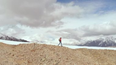 Sırt çantalı, güneş gözlüklü, yürüyüş direkleri olan genç bir adamın dağdan aşağı inişini diğer dağların ve geçitlerin arka planında karın yanında yürürken görüntüsü.