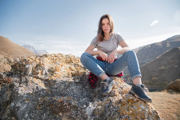Mulher caucasiana jovem e atraente em camiseta cinza e jeans senta-se em um penhasco contra as montanhas em um dia ensolarado — Fotografia de Stock