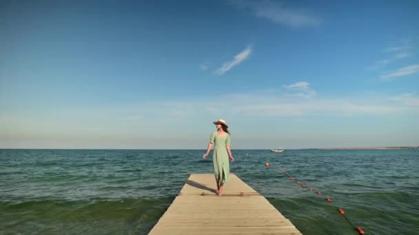 An attractive Caucasian girl in a green summer dress and a straw hat walks along the wooden pier of the sea coast in sunny weather. Rest at the sea. Travel video — Stock Video