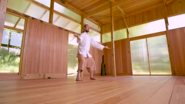 Un hombre caucásico de pelo largo con ropa ligera y suelta practica qigong tai chi en una sala de práctica de madera en verano. Movimientos lentos armonía y calma — Vídeos de Stock