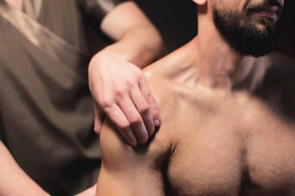Close-up Male shoulder and deltoid muscle massage to athlete muscular man in dark room of spa salon while sitting — Stock Photo, Image