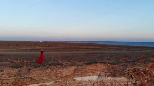 Vista aérea de una joven hermosa mujer en un largo vestido rojo en una roca de montaña mirando al mar, una joven con un vestido rojo con el pelo revoloteando en el viento en el mar en la roca — Vídeos de Stock