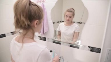 Banyoda aynanın önünde duran genç bir kadın diş fırçasına diş macunu sürüyor.