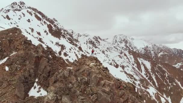 A sporty man in an orange winter suit stands alone in the snow in the evening against the backdrop of epic snow-capped mountains and peaks. Aerial view