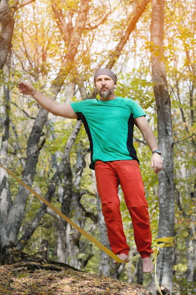 Um homem barbudo adulto que se equilibra em um slackline apertado está envolvido no ato de equilíbrio na floresta de outono — Fotografia de Stock