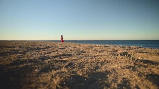 Romantyczna kobieta w czerwonej sukience stoi na wybrzeżu. Lekka sukienka trzepocze na wietrze. Trzyma słomkowy kapelusz. — Wideo stockowe