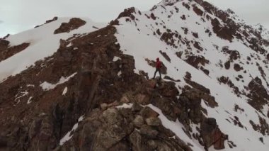 Turuncu kışlık takım elbiseli sportif bir adam akşam vakti kar altında destansı kar örtülü dağların ve zirvelerin zemininde tek başına duruyor. Hava görünümü