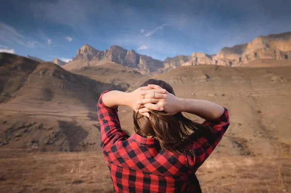 Fiatal, boldog nő pólóban és farmerben magasan áll a hegyekben kinyújtott karokkal, élvezve a hegyekre néző kilátást és a táj epikus természetét. — Stock Fotó