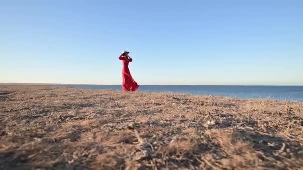 Seorang wanita romantis dengan gaun merah berdiri di pantai tinggi. Sebuah gaun cahaya berkibar-kibar dalam angin. Berpegangan pada topi jerami — Stok Video