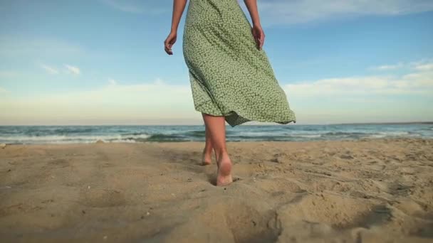 Ноги молодої жінки в повільному русі ходять босоніж на пляжі. Залишивши сліди в піску Турист у відпустці біля моря. Жінка в красивій сукні на вітрі — стокове відео