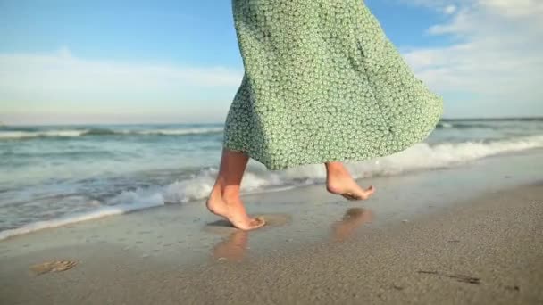 Piernas de una joven en cámara lenta caminando descalza por la playa. Dejando huellas en la arena Turista de vacaciones junto al mar. Mujer en un hermoso vestido revoloteando en el viento — Vídeo de stock