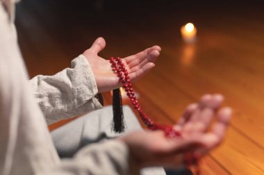Meditasyon yapan bir keşişin erkek eli, mum ışığında karanlık bir odada kırmızı bir tespih tutar. Pratik ve aydınlanma