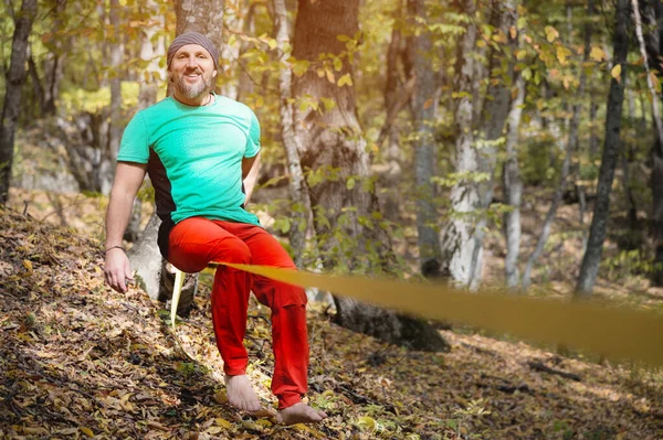 Um homem barbudo em equilíbrio de idade, sentado em uma tensa slackline na floresta de outono. Lazer ao ar livre — Fotografia de Stock