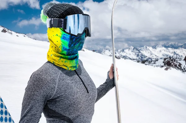 Eine attraktive, schlanke Skifahrerin steht ohne Jacke hoch in den Bergen und hält ihre Skier vor dem Hintergrund hoher schneebedeckter Gipfel und Wolken. — Stockfoto