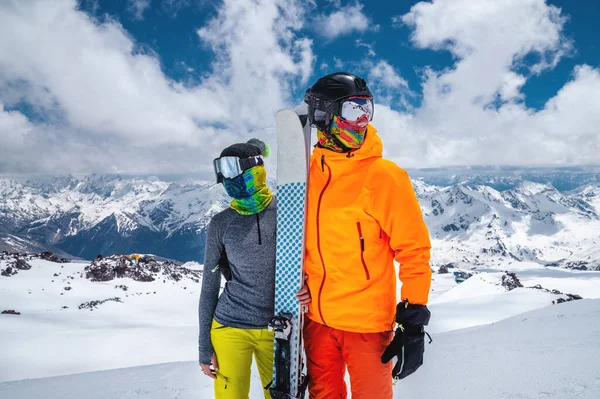 Um jovem casal apaixonado, um homem e uma mulher em trajes de esqui, ficam ao lado de esquis em um obmnik contra o fundo de montanhas cobertas de neve. Amor de atletas — Fotografia de Stock