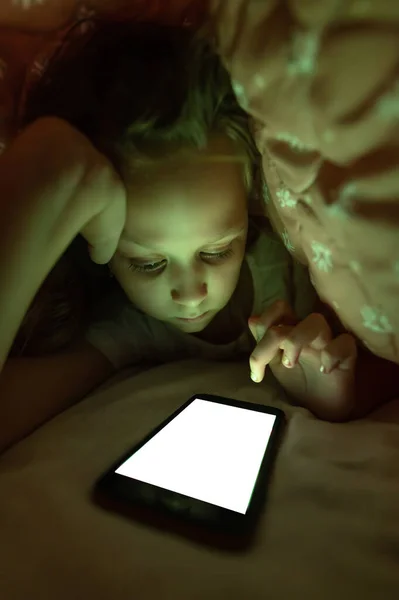 Молода дівчина під обкладинками. граючи з мобільними телефонами і перевіряючи електронну пошту в спальні в квартирі. Концепція дитячої залежності від гаджетів — стокове фото