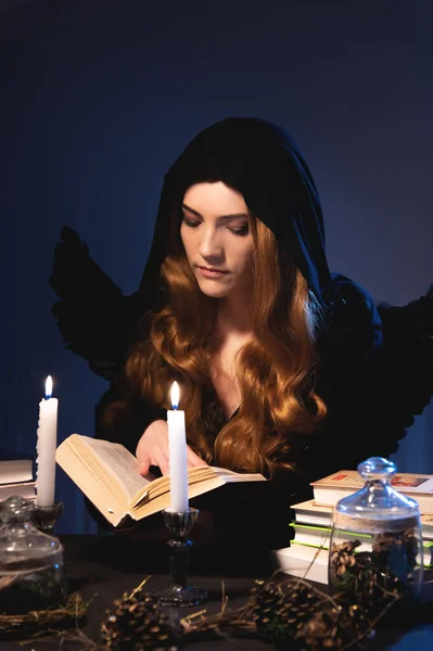Una hechicera con capucha negra halaga un libro rodeado de velas y accesorios de brujería — Foto de Stock