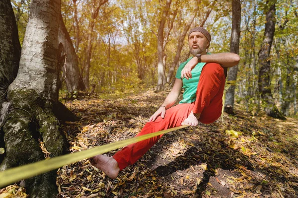Um homem barbudo em equilíbrio de idade, sentado em uma tensa slackline na floresta de outono. Lazer ao ar livre — Fotografia de Stock