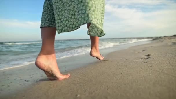 Piernas de una joven en cámara lenta caminando descalza por la playa. Dejando huellas en la arena Turista de vacaciones junto al mar. Mujer en un hermoso vestido revoloteando en el viento — Vídeos de Stock