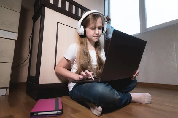 Een slim meisje van zeven met een witte koptelefoon met een laptop in haar handen duwt op de vloer in haar kamer. De jonge generatie op het internet en IT-technologie — Stockfoto