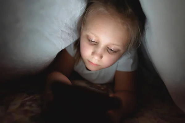 Küçük bir kız elinde akıllı telefon cihazıyla gece bir battaniyenin altında. Çocuklar için internet ve oyun bağımlılığı. Uykusuzluk — Stok fotoğraf