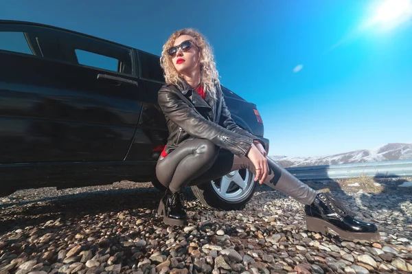 Une jolie blonde en lunettes de soleil et vêtements en cuir se trouve près d'une voiture de sport noire sur une route de campagne dans les montagnes au coucher du soleil. Style rétro années 90 — Photo