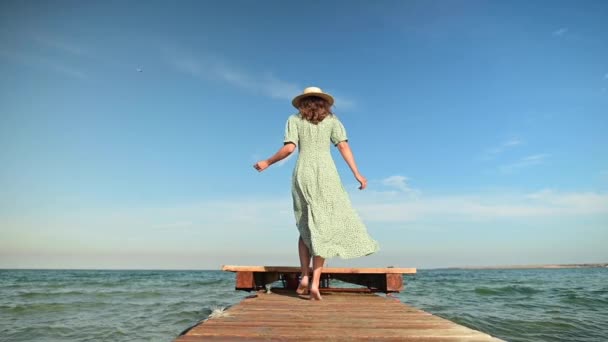 Une jeune femme vêtue d'une robe d'été verte et d'un chapeau de paille au ralenti se promène le long d'une jetée en bois le matin sur la mer pendant le vent. Promenade grâce beauté et féminité — Video