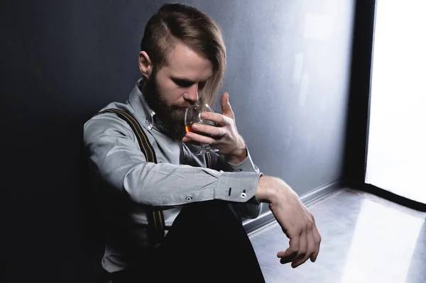 Hombre barbudo deprimido solitario con corte de pelo elegante bebe whisky mientras está sentado junto a la pared en el estudio — Foto de Stock