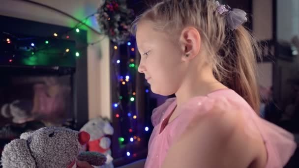 Klein eenzaam vrolijk vet meisje spelen met een zacht speelgoed rat op de achtergrond van een elektrische open haard en knipperen nieuwjaar lichten op kerstavond — Stockvideo